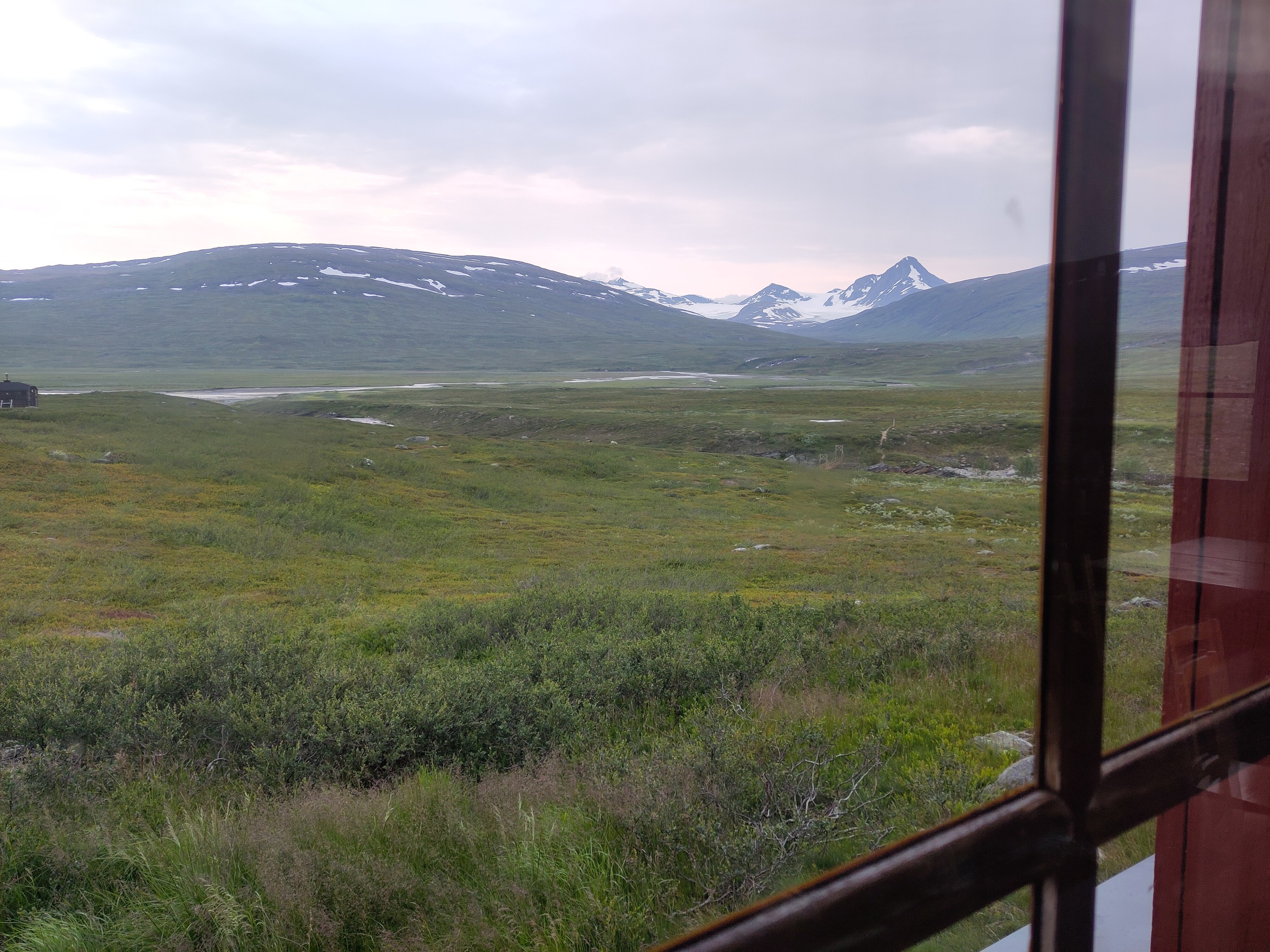 A view out a window with fairly flat rolling landscape in the foreground gives way to dark peaks between white glaciers in the distance.