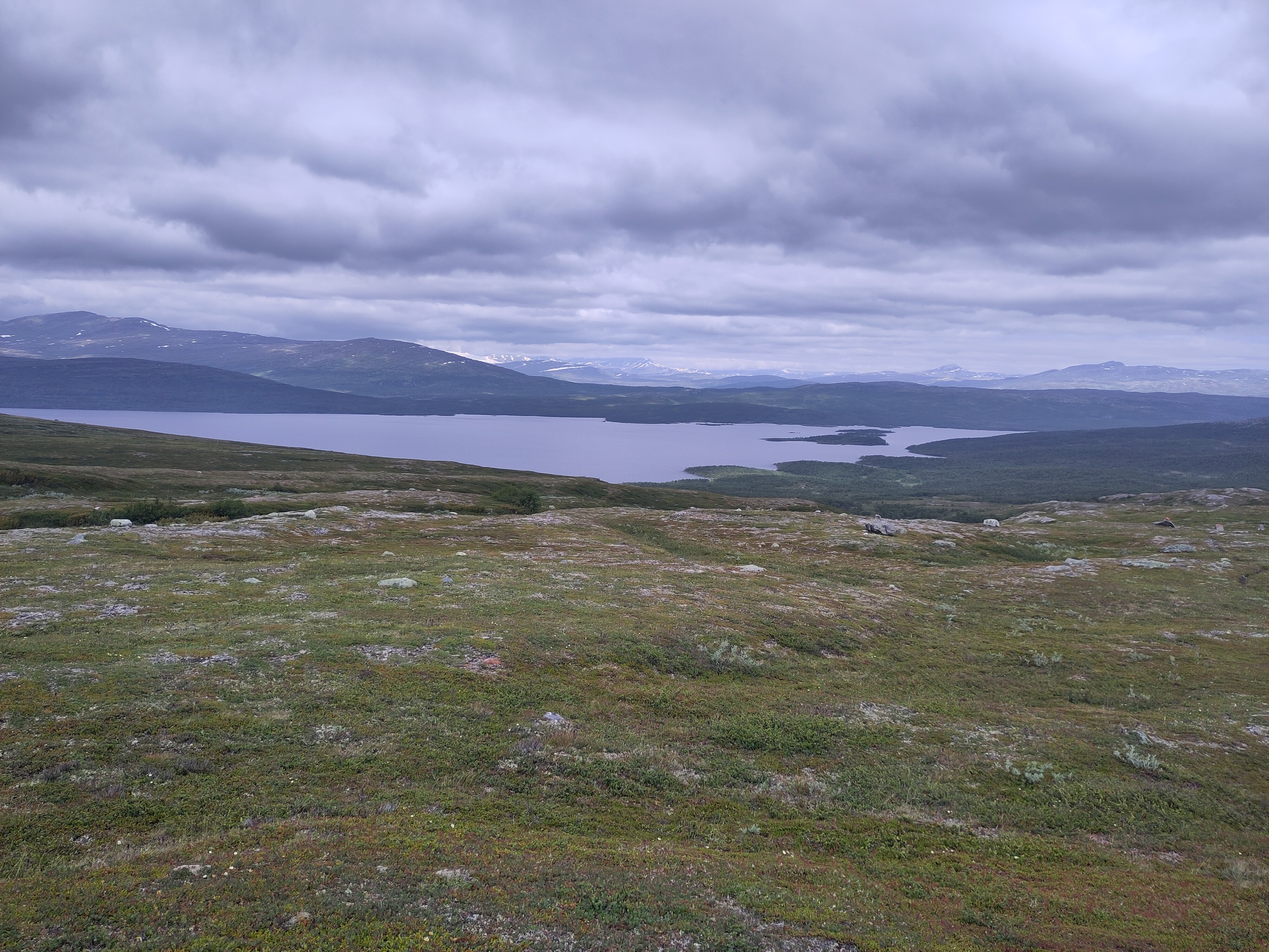 A view of lake Mavas from the slope on the southern side. It stretches out both west and east from the photo, even though it is from far away. There are some islands to the right on the frame.The photo is taken from above the tree line, but you can tell there is lots of woods on both sides of the lake. Above, a cloudy sky, and you can maybe make out some mountains in the distance.
