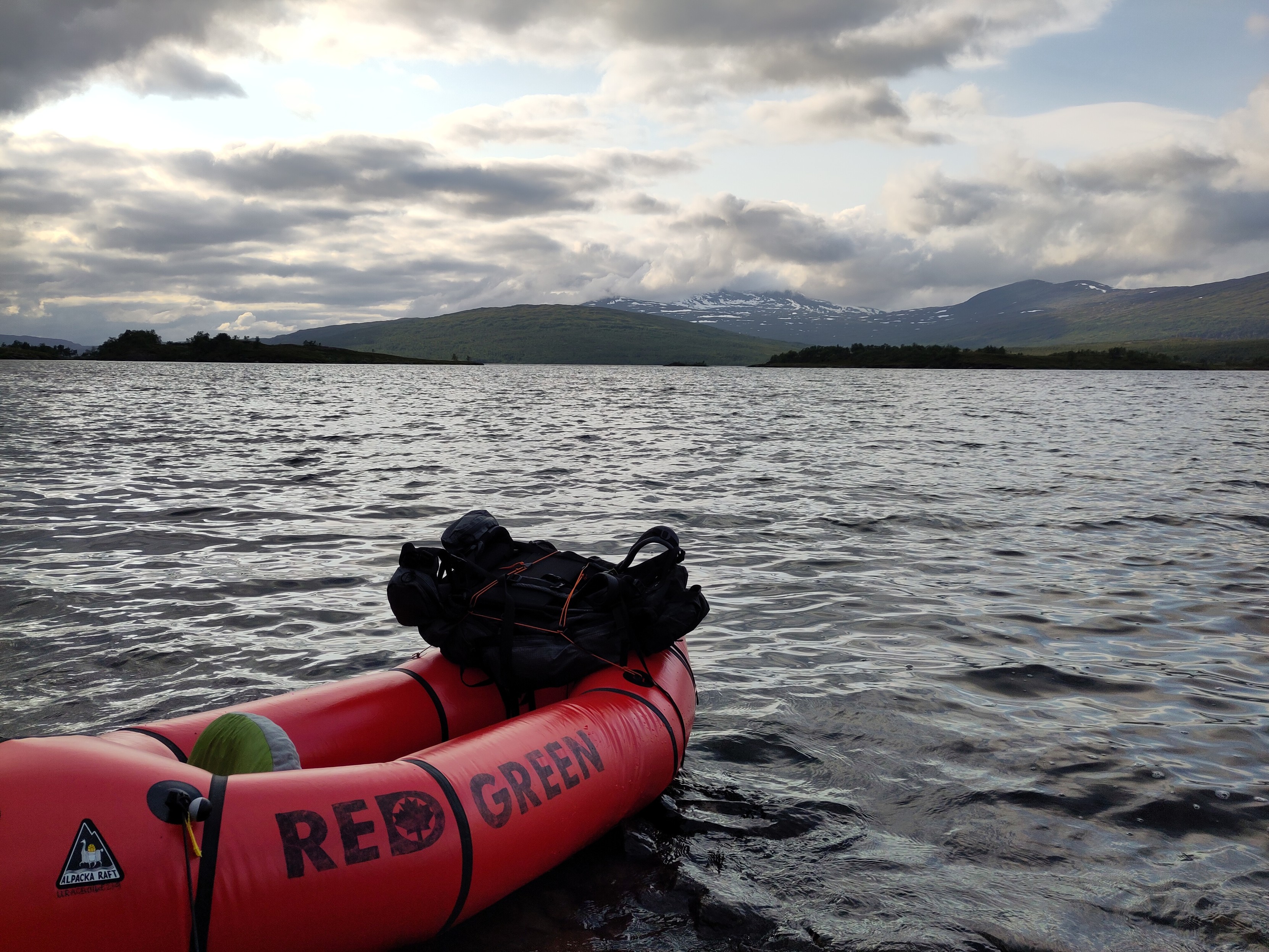 The red packraft labelled "Red Green" with a backpack strapped to the bow, looks ready for departure from the shore. The sky has started to break up a bit, showing some spots of blue sky.