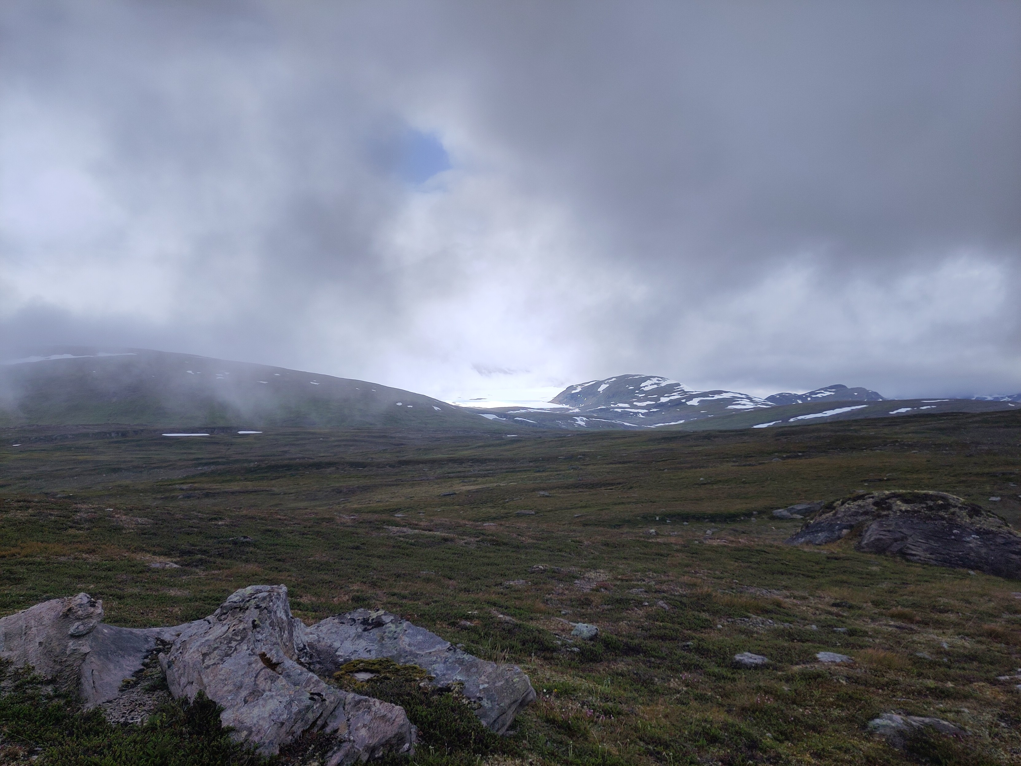 A view from afar of a glacier between a couple of mountains glowing white in sudden sunlight, when the cloud cover is almost complete.

In the foreground is a wide expanse of small ridges and mostly flat ground.