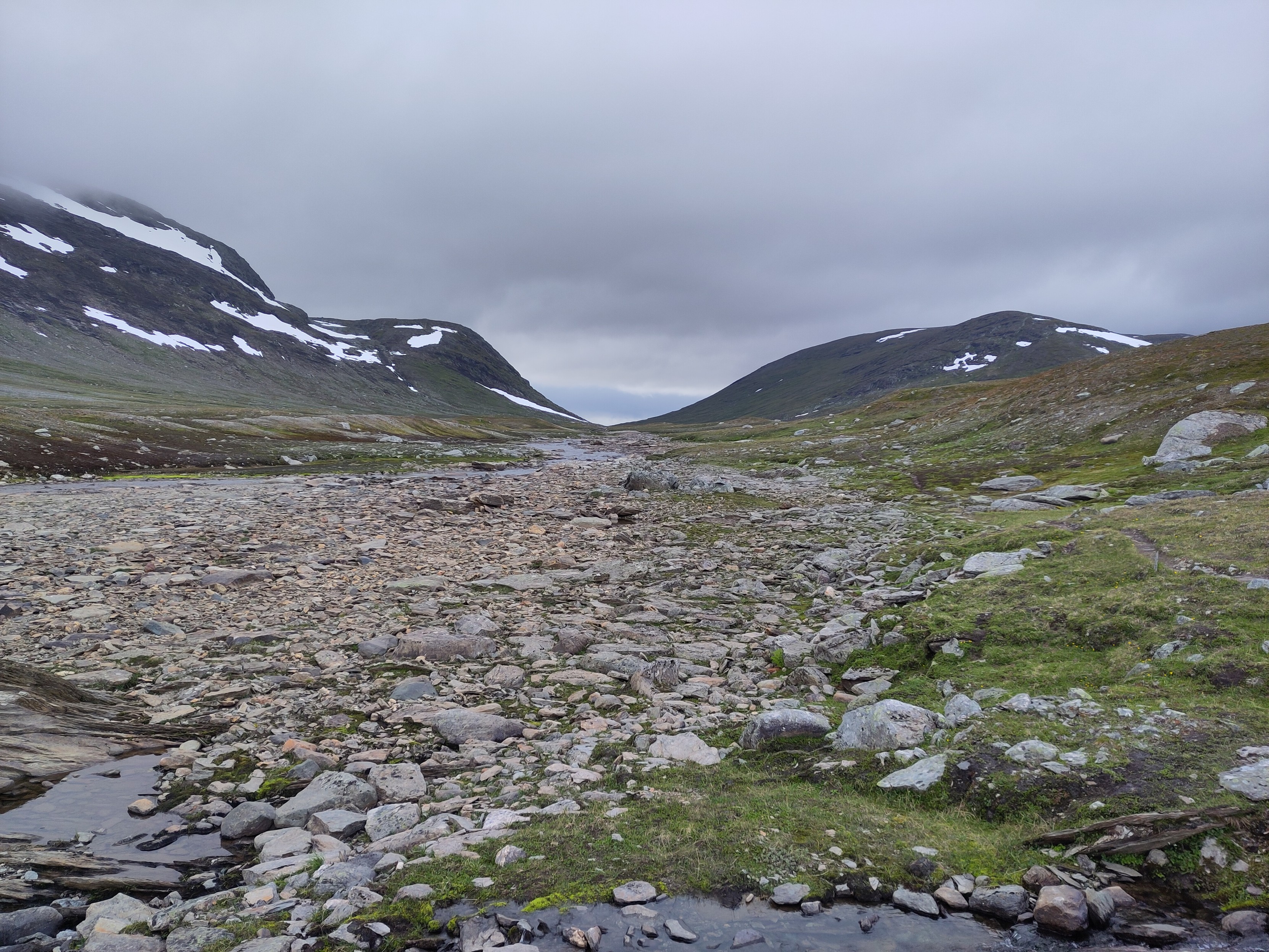 A wide rocky stream bed with a stream on the other side from the photographer. The stream comes down from a long valley where the mountains on either side are dark with plenty of white snow fields.