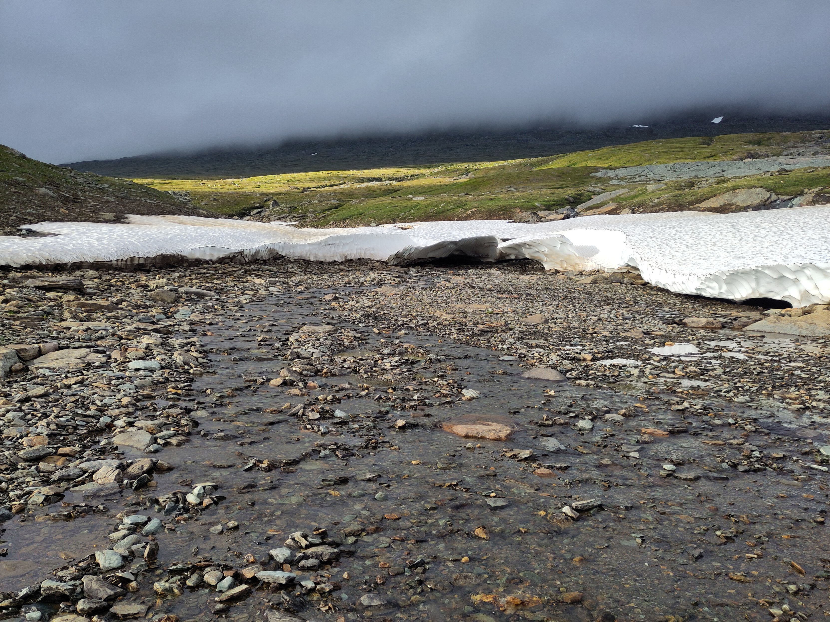 A very shallow stream coming out of a snow field upstreams. The stream bed is rocky and the sky is cloudy, yet above the stream a yellow-green section of land is lit by the sun.