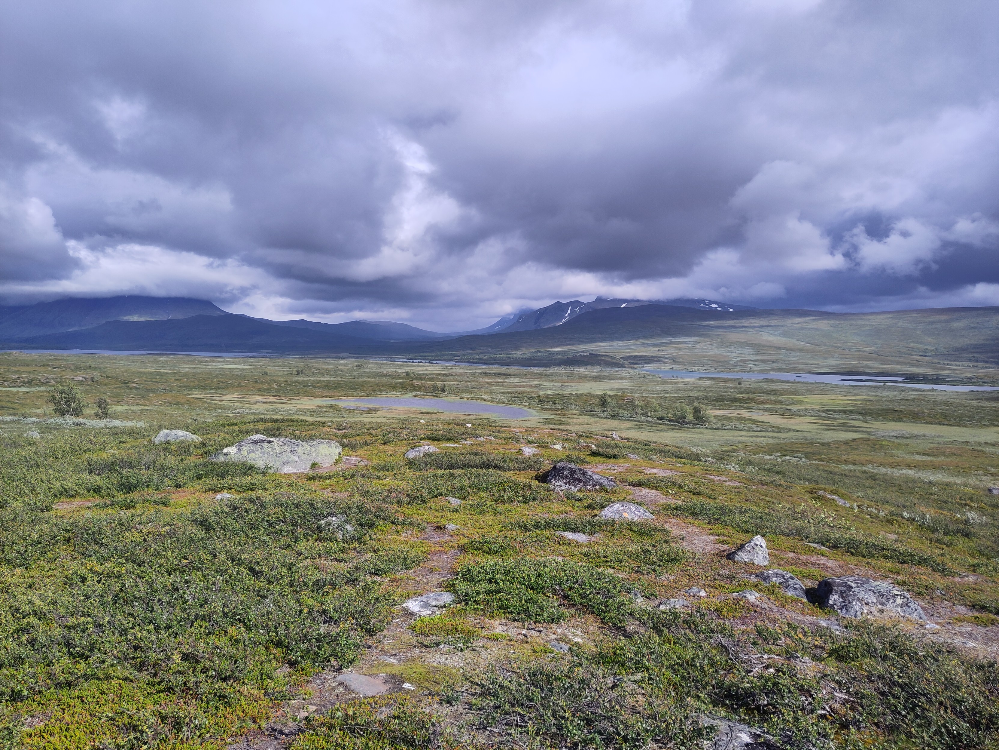 A view towards Akka and Sarek, with dark peaks disappearing up into low clouds. In the foreground a fairly flat and green landscape slopes down.