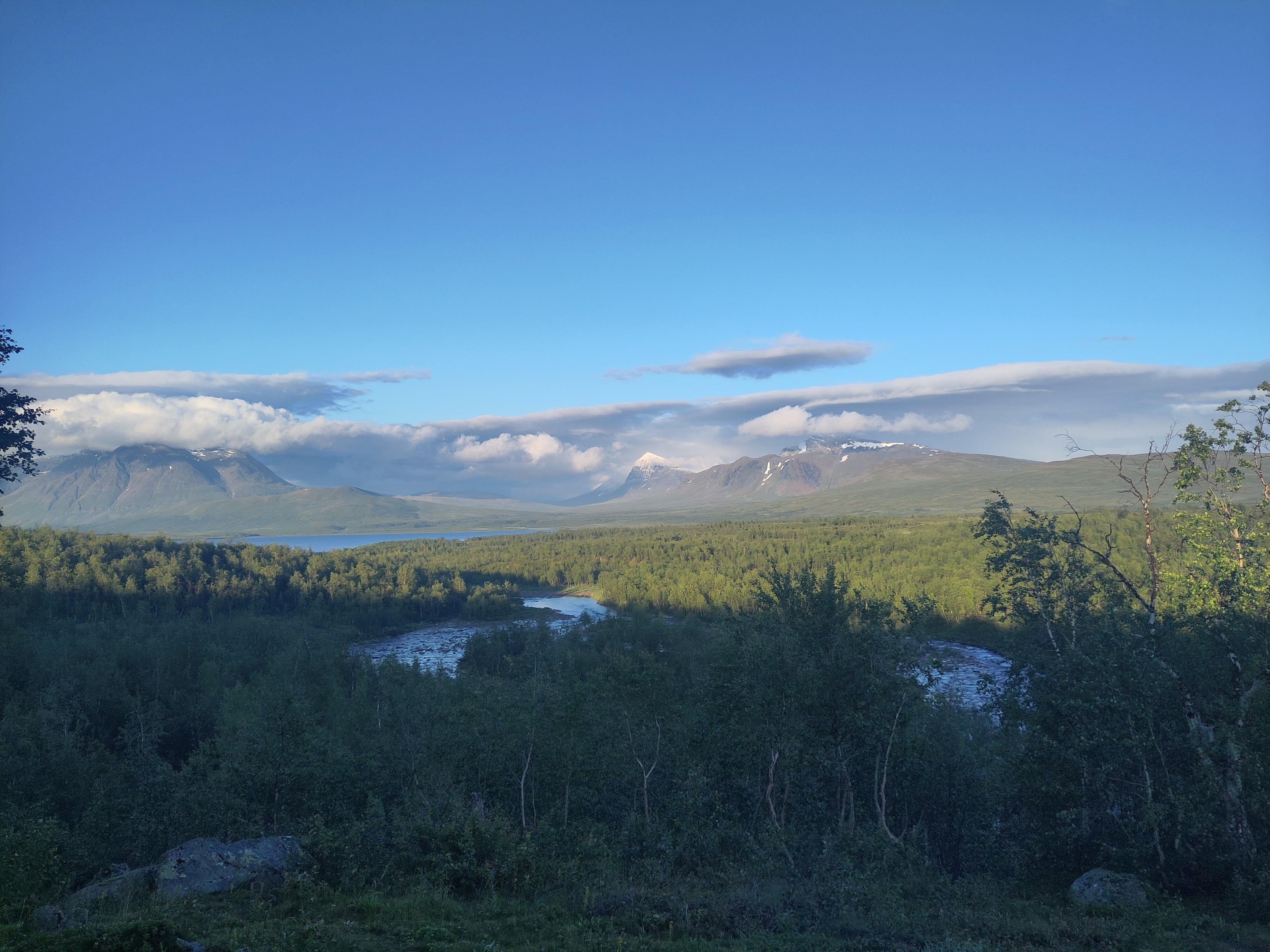 Akka and Gisuris and more remote Sarek peaks with a fresh white dusting at higher altitudes. There are clouds around the mountains in the distance, but most of the sky is blue and most of the land before the mountains are sunlit and green
