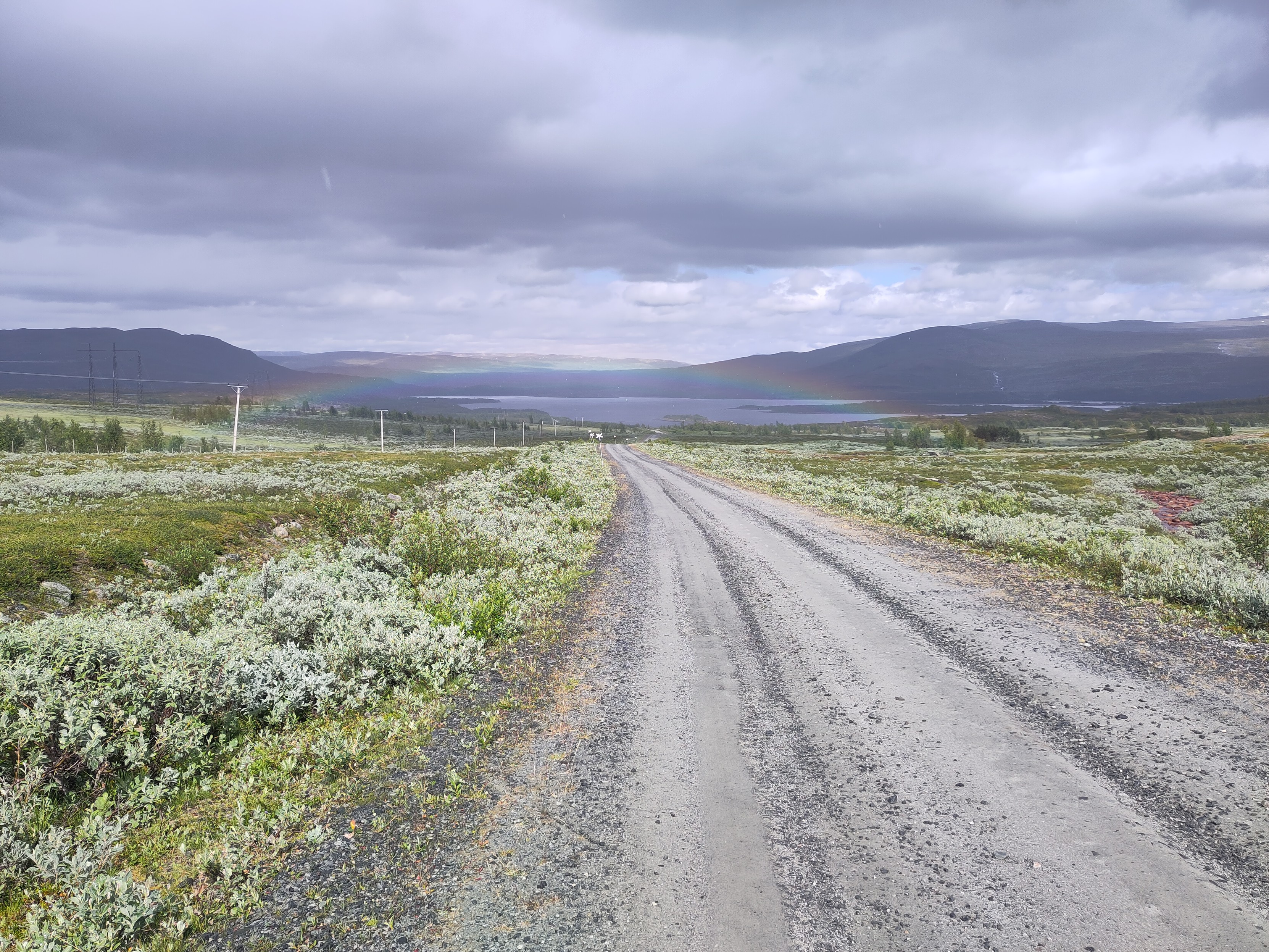 A gravel road up above the tree line, with a lake straight ahead. Above the lake there is a rainbow.