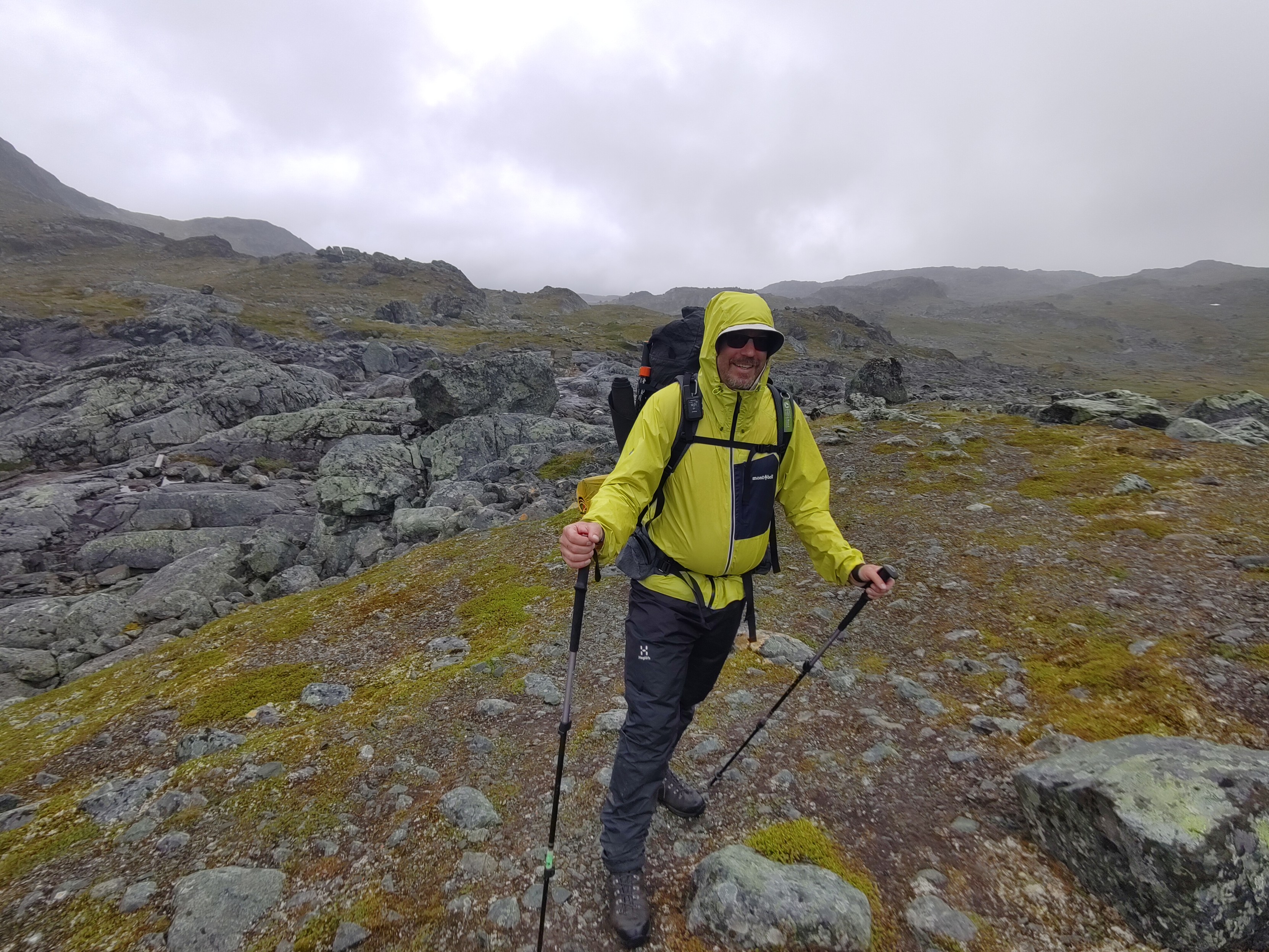 A white dude in yellow rain coat standing in a broken landscape with some light mists in the distance.
