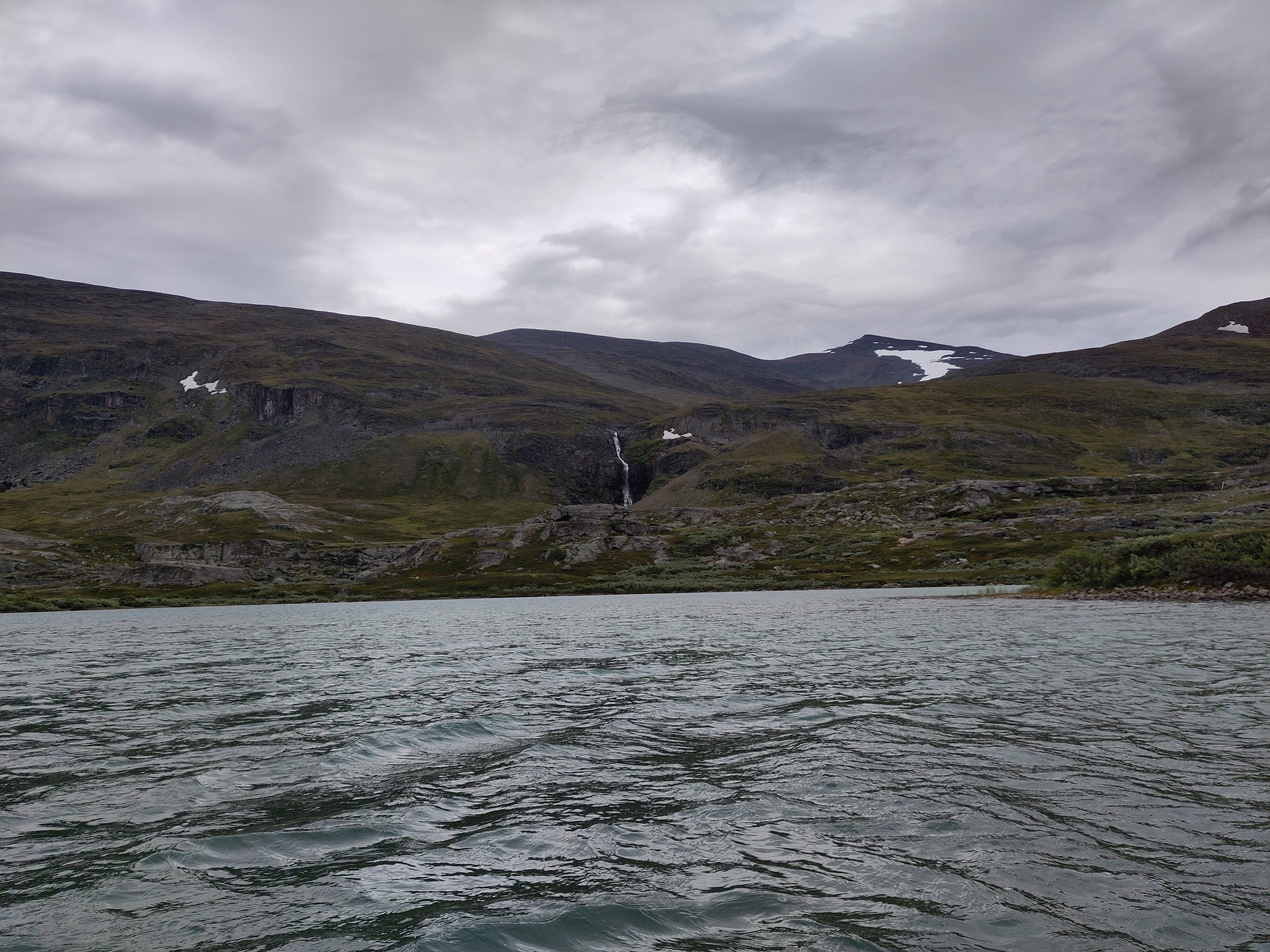 A steep part of a mountain side with a tall waterfall, as seen from a lake.
