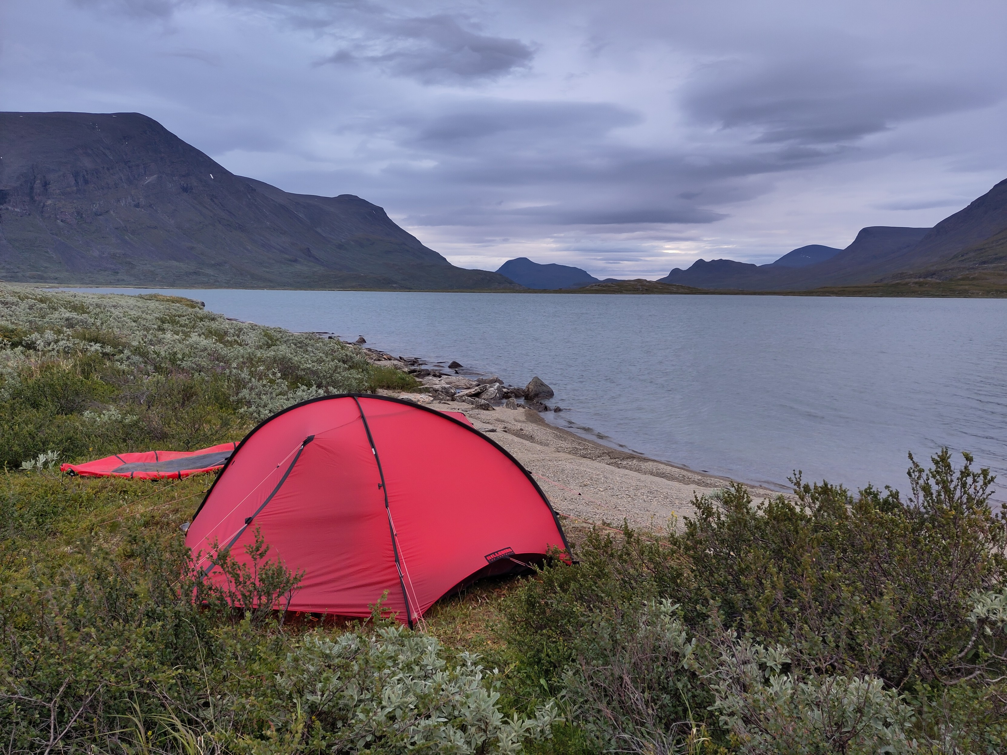 A red tent and behind it a deflated packraft, setup next to a little beach by a lake. In the distance more mountains and a cloudy sky.